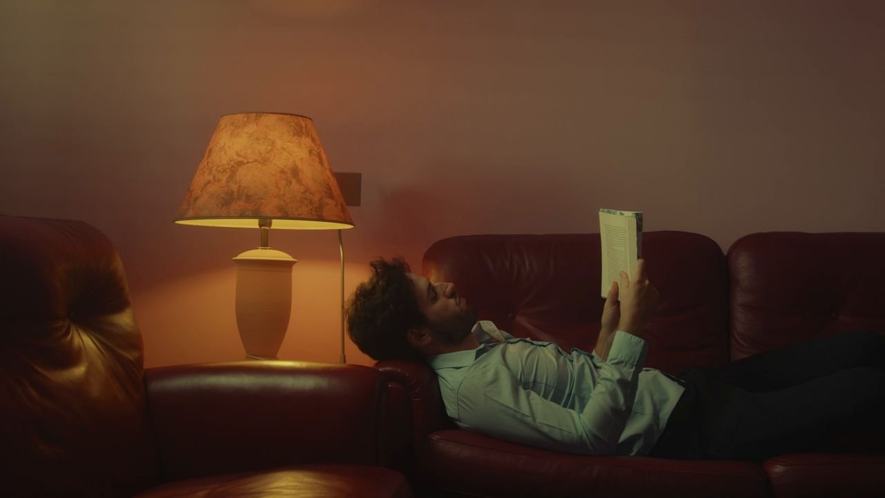 Production still: a man reading on a sofa under a glowing table lamp